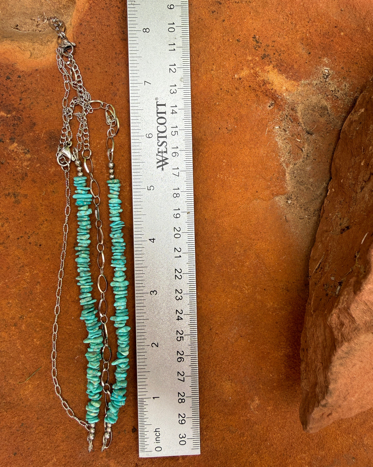 A silver chain necklace with turquoise blue beads and small white stones, displayed on a terracotta surface next to a metal ruler for scale.