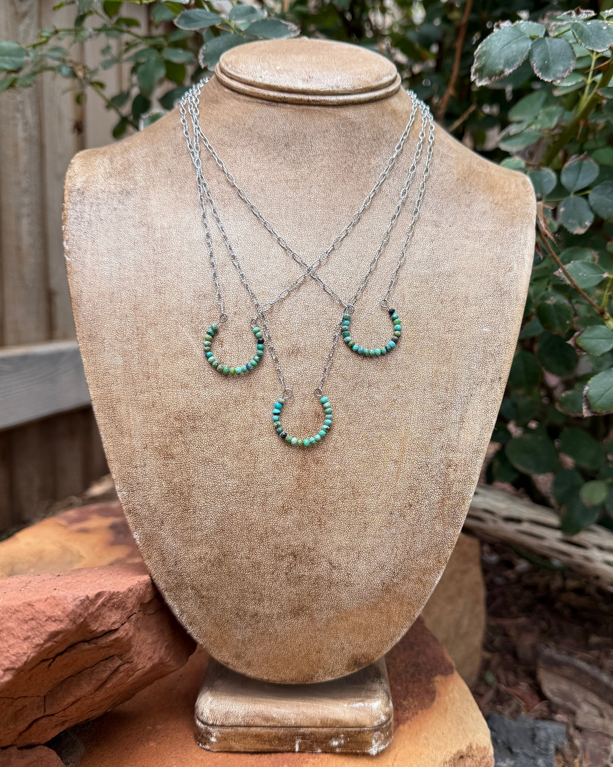 A three-layer silver necklace with turquoise horseshoe beaded pendant displayed on a beige jewelry bust outdoors.