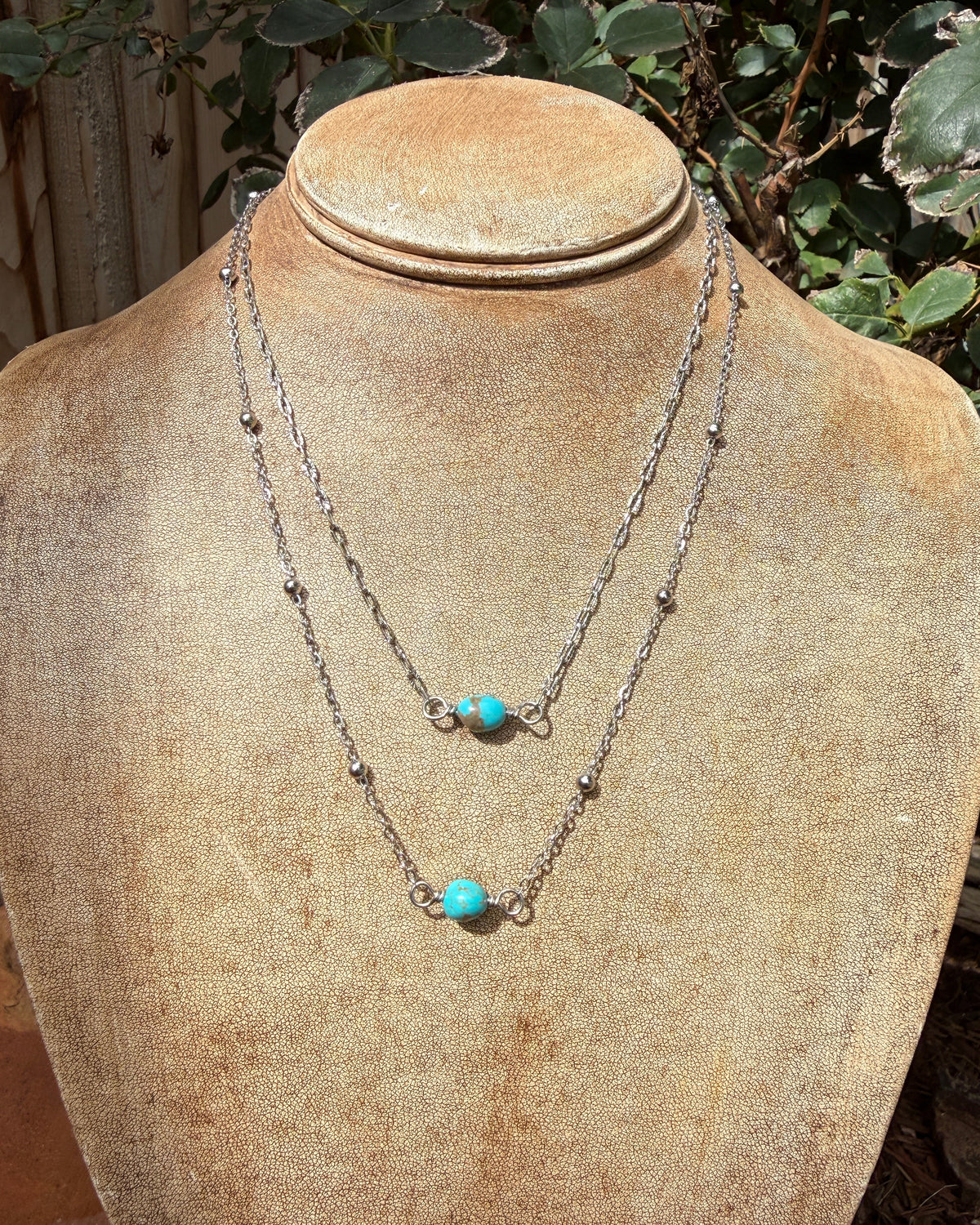 Two layered silver chain necklaces with small bead details and turquoise stone accents, displayed on a tan jewelry bust outdoors with green foliage in the background.