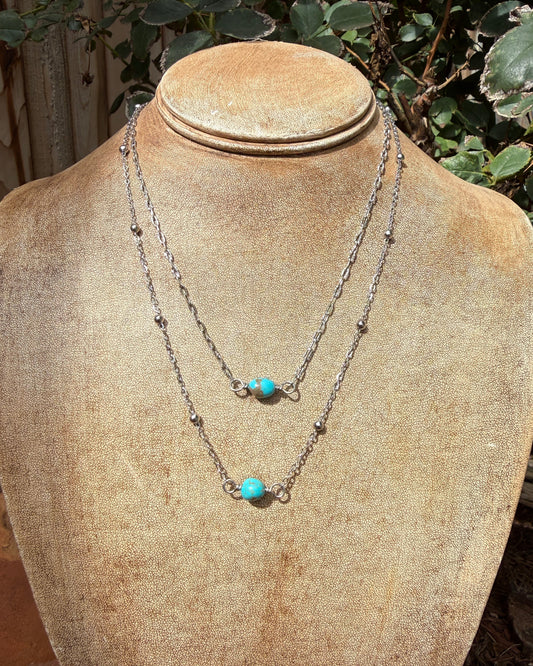 Two layered silver chain necklaces with small bead details and turquoise stone accents, displayed on a tan jewelry bust outdoors with green foliage in the background.