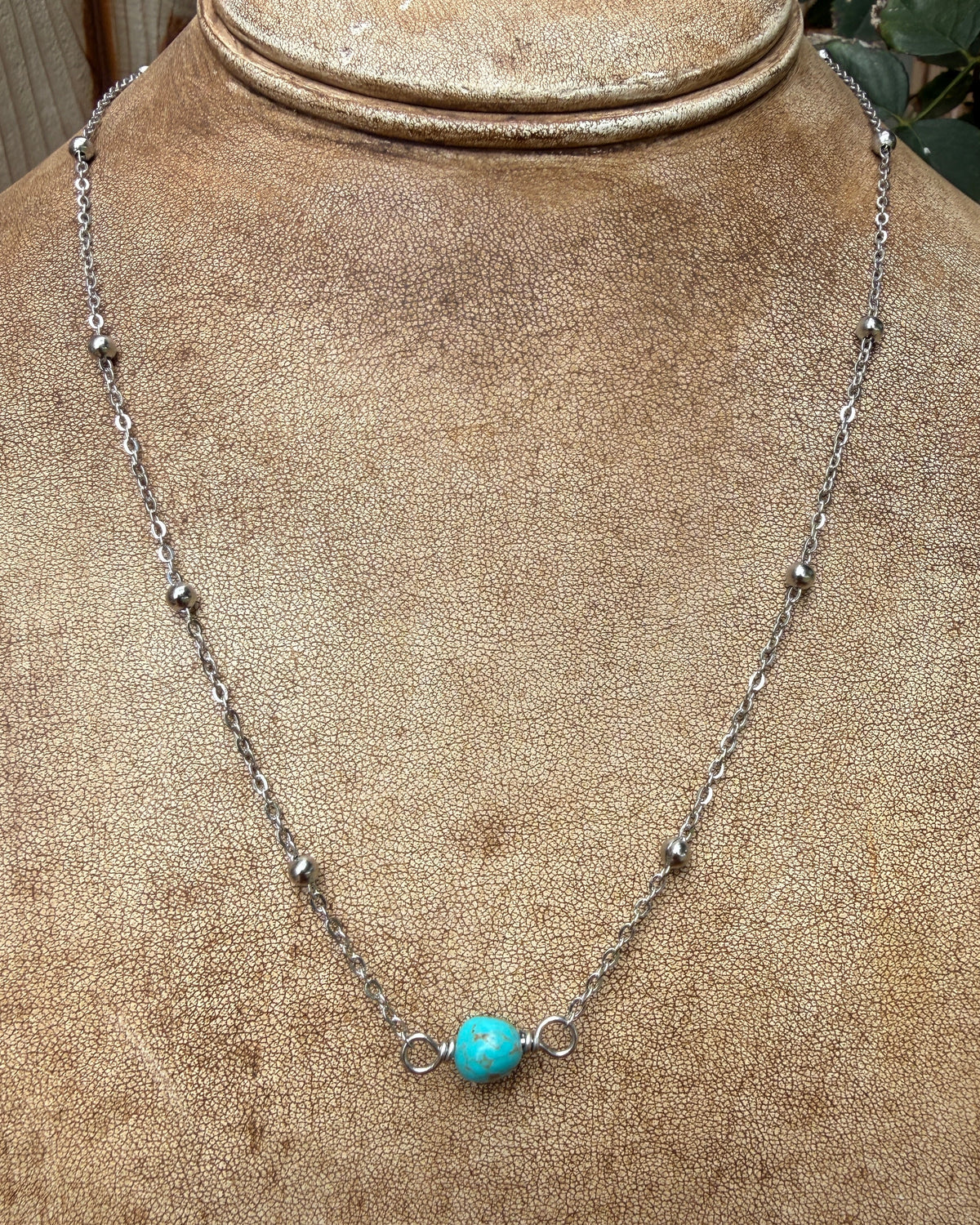 A silver-toned chain necklace with a single round turquoise bead pendant, displayed on a textured tan jewelry bust.