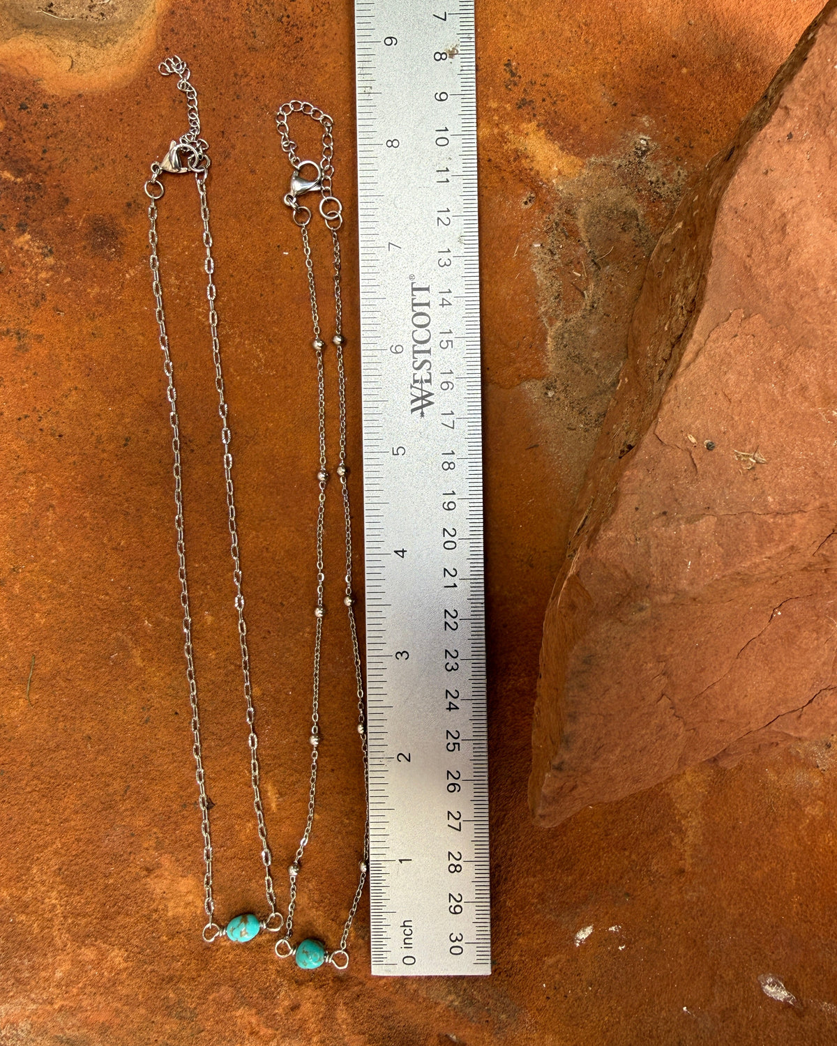 A silver chain necklace with two small turquoise bead pendants is displayed next to a metal ruler for scale on a textured orange-brown stone surface.