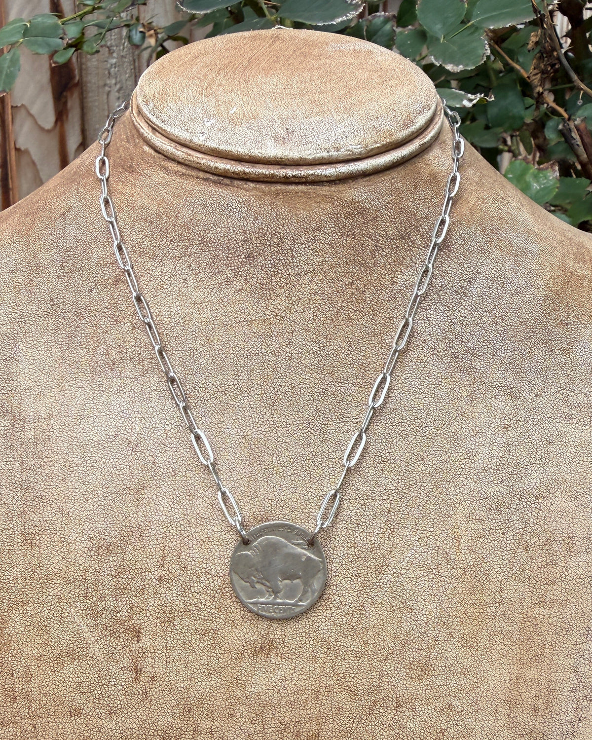 A silver chain necklace with a circular coin-style pendant featuring an animal design, displayed on a textured tan jewelry bust outdoors with green foliage and a wooden fence in the background.