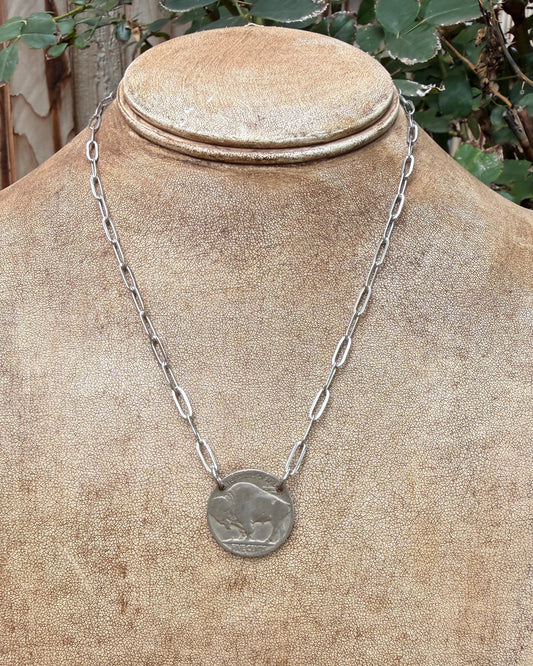 A silver chain necklace with a circular coin-style pendant featuring an animal design, displayed on a textured tan jewelry bust outdoors with green foliage and a wooden fence in the background.