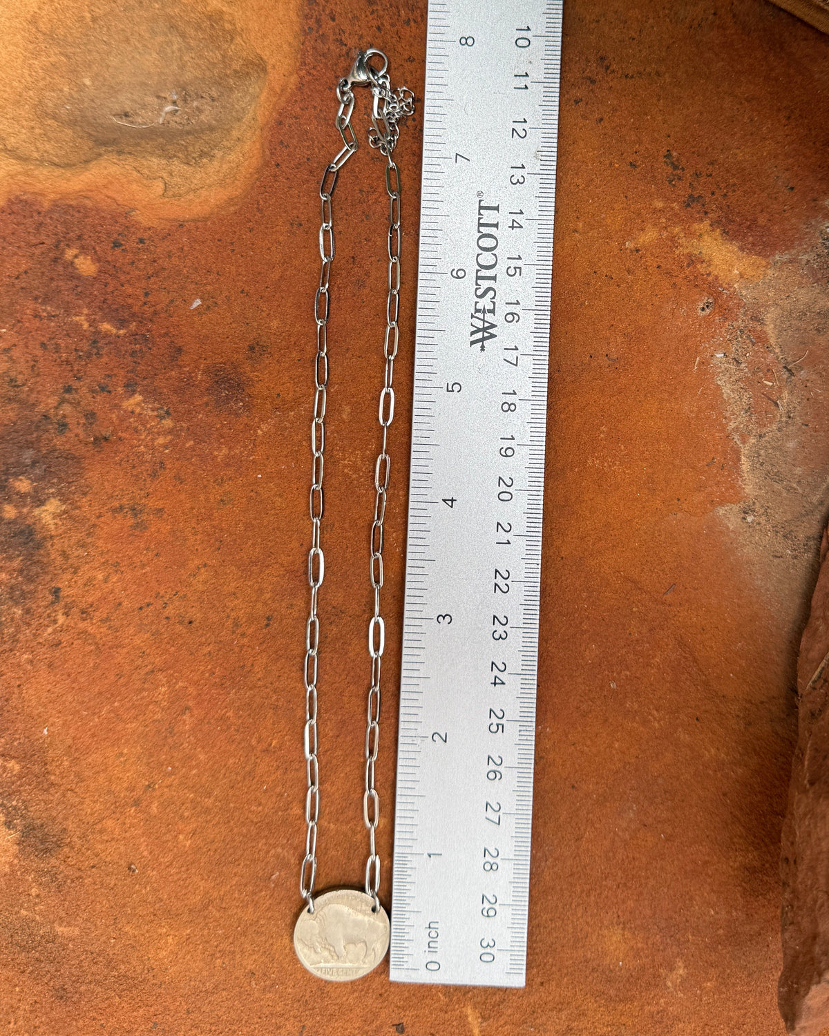 A silver-toned chain necklace with a small round pendant lies next to a white measuring ruler on a textured orange-brown surface.