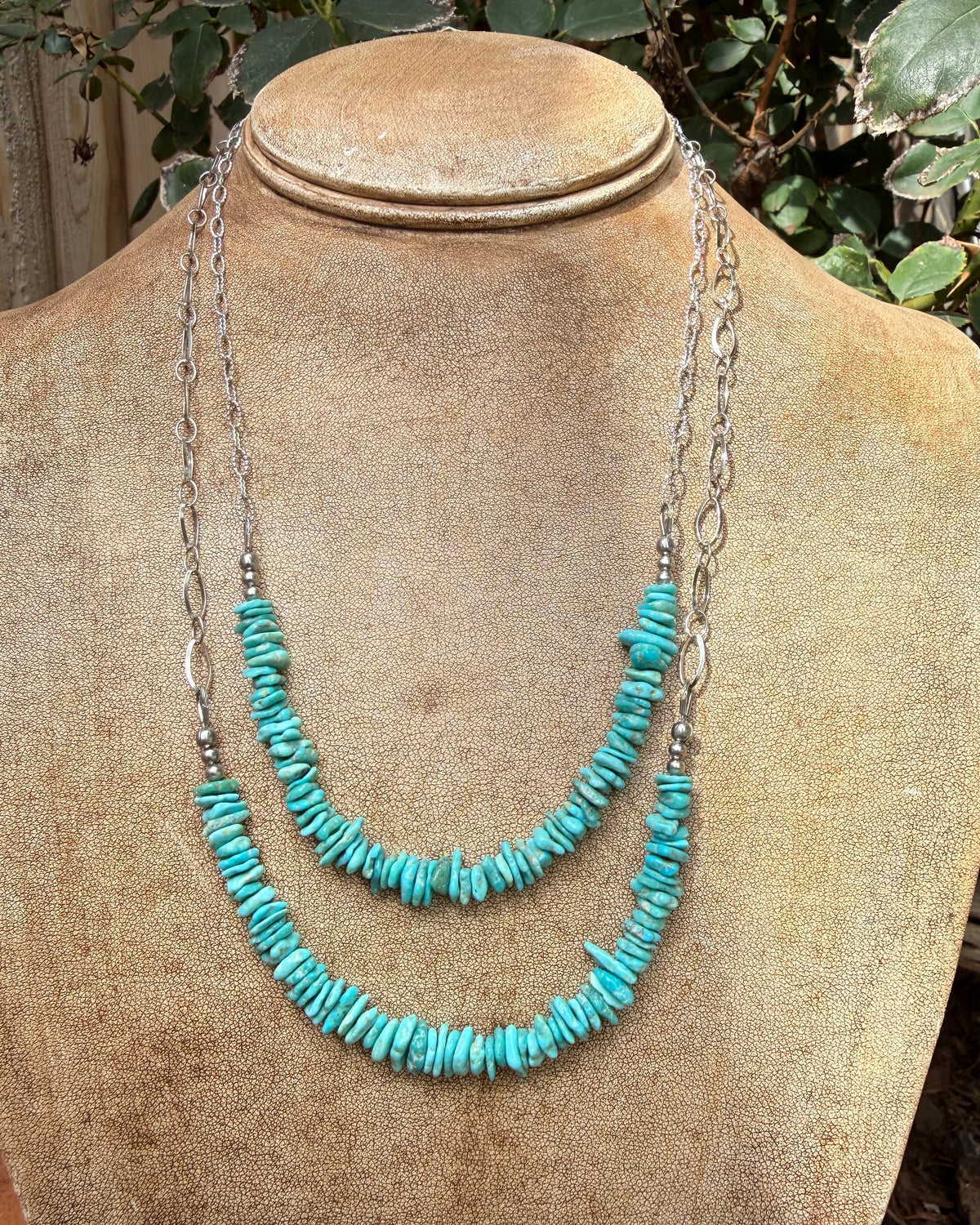 A two-strand necklace displayed on a tan fabric mannequin bust, featuring turquoise-colored stone chips on silver chains, set against a background of green foliage.