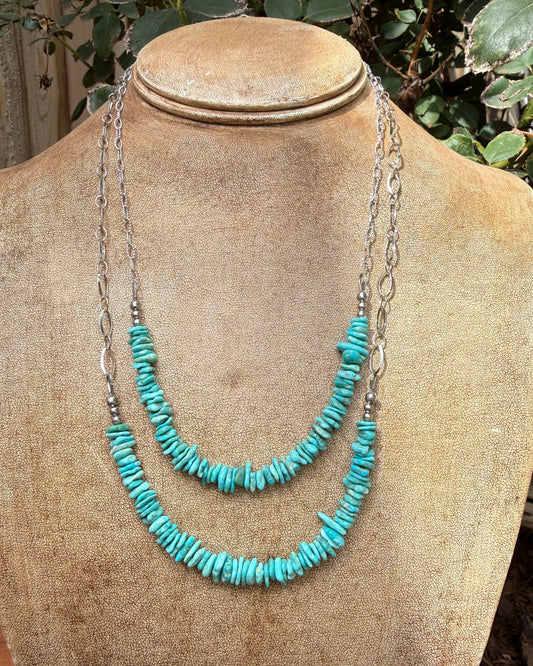 A two-strand necklace displayed on a tan fabric mannequin bust, featuring turquoise-colored stone chips on silver chains, set against a background of green foliage.