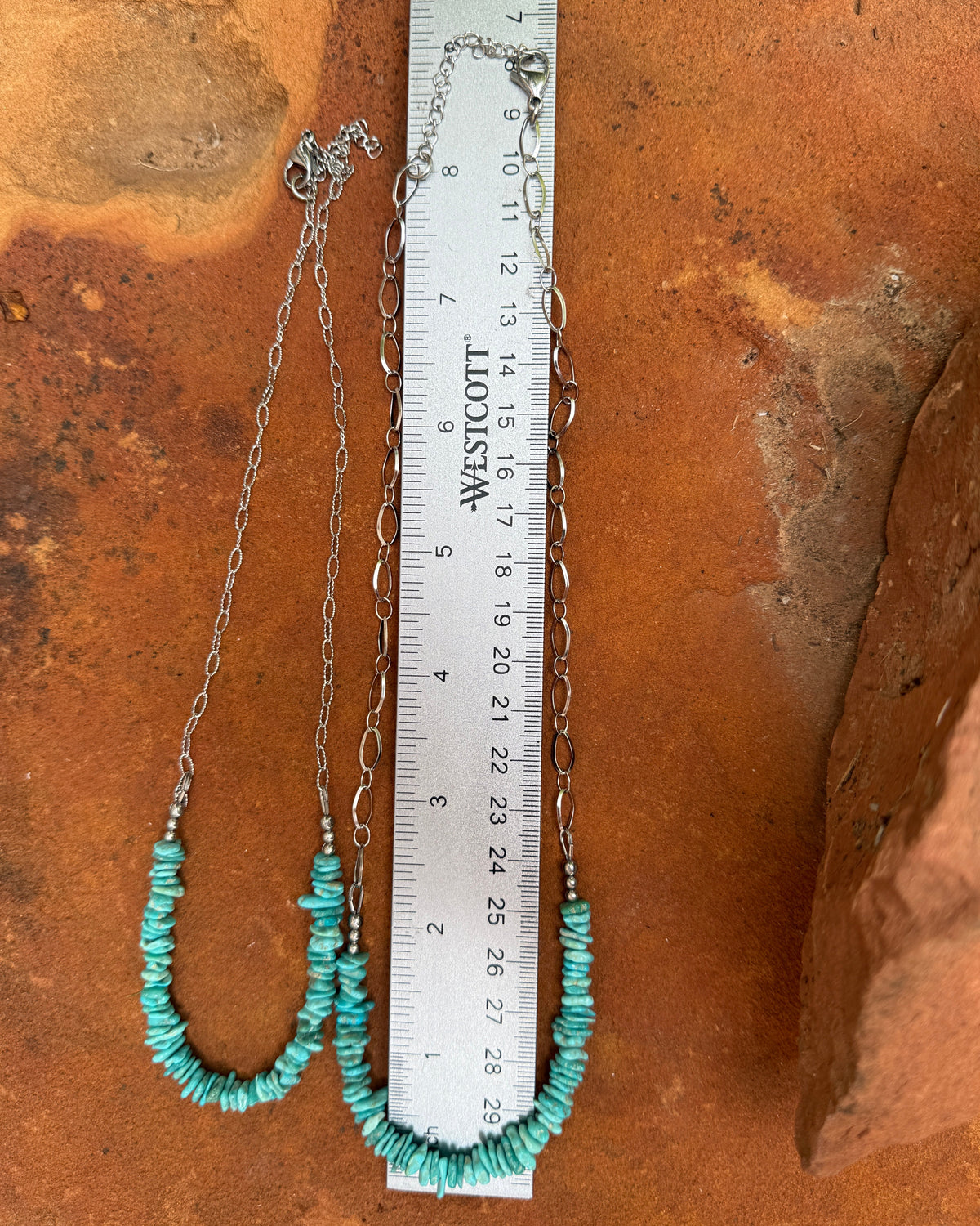 A handmade turquoise and silver necklace laid out on a terracotta surface next to a metal ruler for scale.