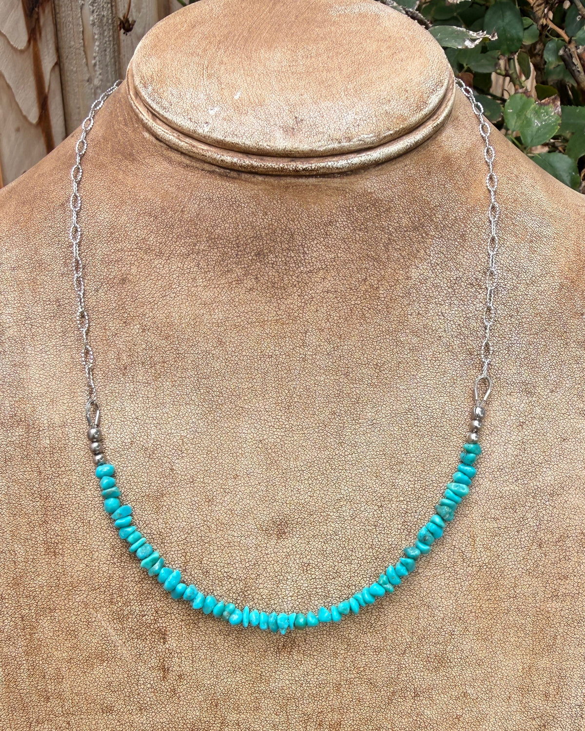 A turquoise beaded necklace with a silver chain, displayed on a tan textured jewelry bust. The background shows a rustic outdoor setting with a wooden fence and green foliage.