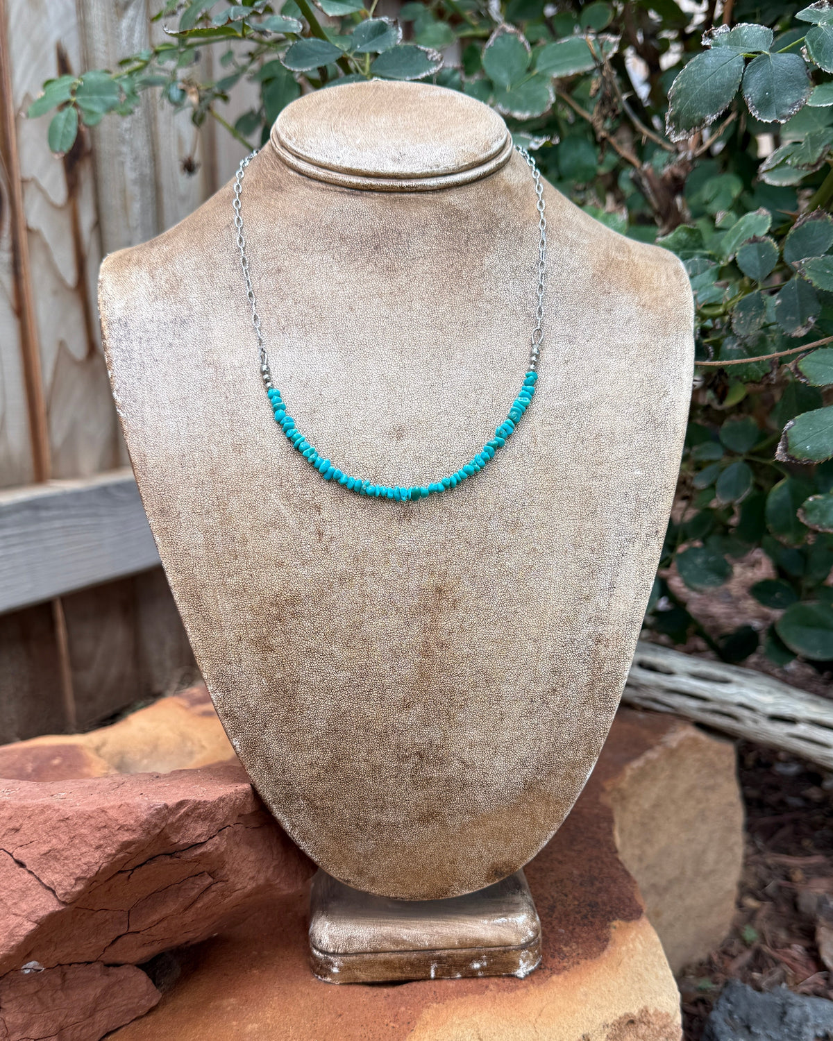 A turquoise beaded necklace on a silver chain, displayed on a stone bust mannequin outdoors on a rock with garden plants and a wooden fence in the background.