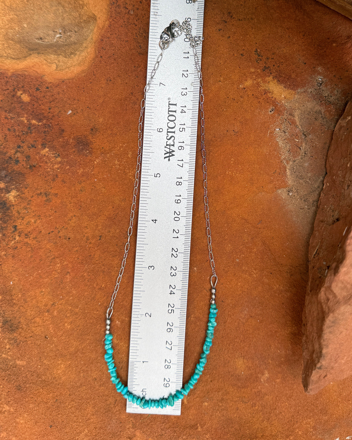 A silver-toned beaded necklace with turquoise blue beads and small red spacer beads, laid out on a metal ruler over a terracotta surface with rocks in the background.