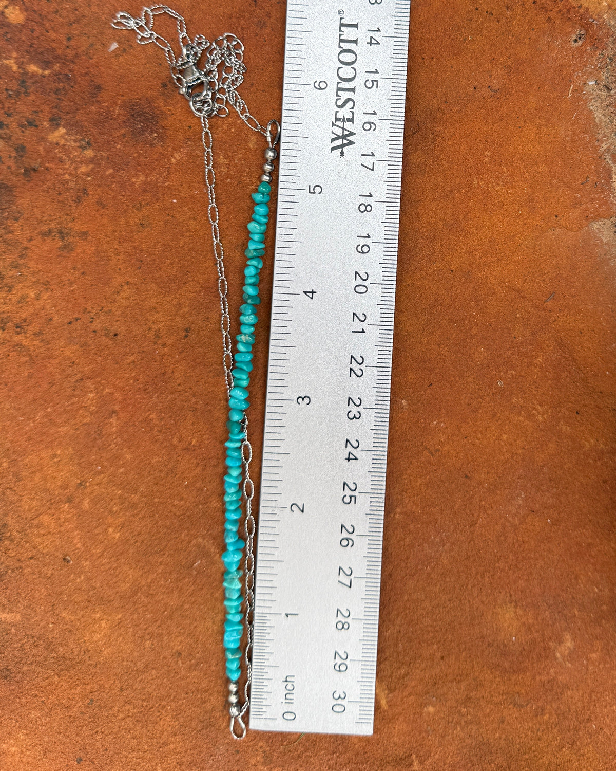 A silver-toned beaded necklace with turquoise blue beads lies on a rusted metal surface next to a metal ruler for scale, with a brick in the upper background.