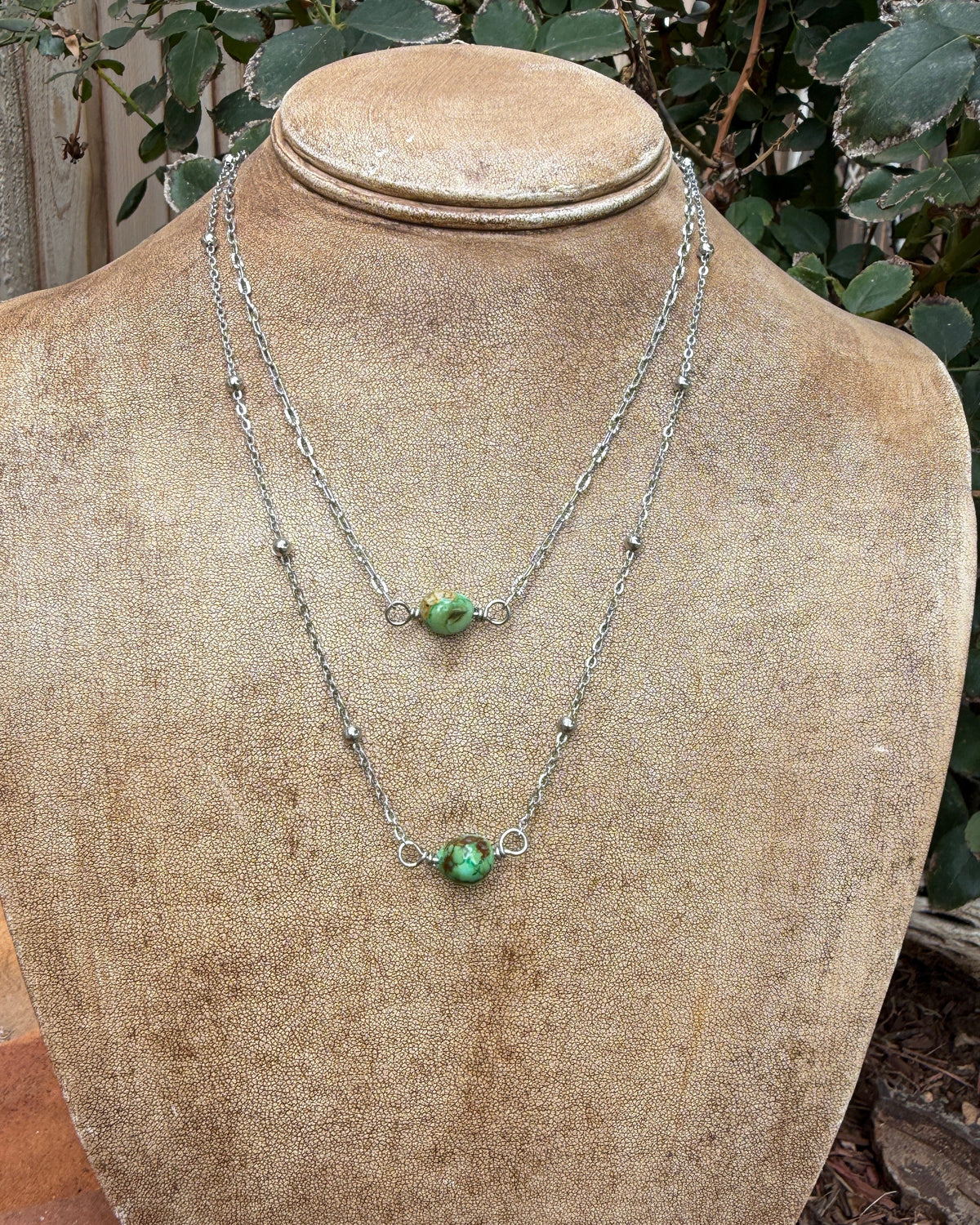 Two layered silver chain necklaces with small green turquoise bead pendants displayed on a tan fabric-covered jewelry bust outdoors, with foliage and garden elements in the background.
