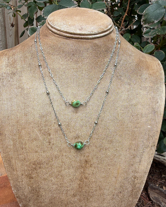 Two layered silver chain necklaces with small green turquoise bead pendants displayed on a tan fabric-covered jewelry bust outdoors, with foliage and garden elements in the background.