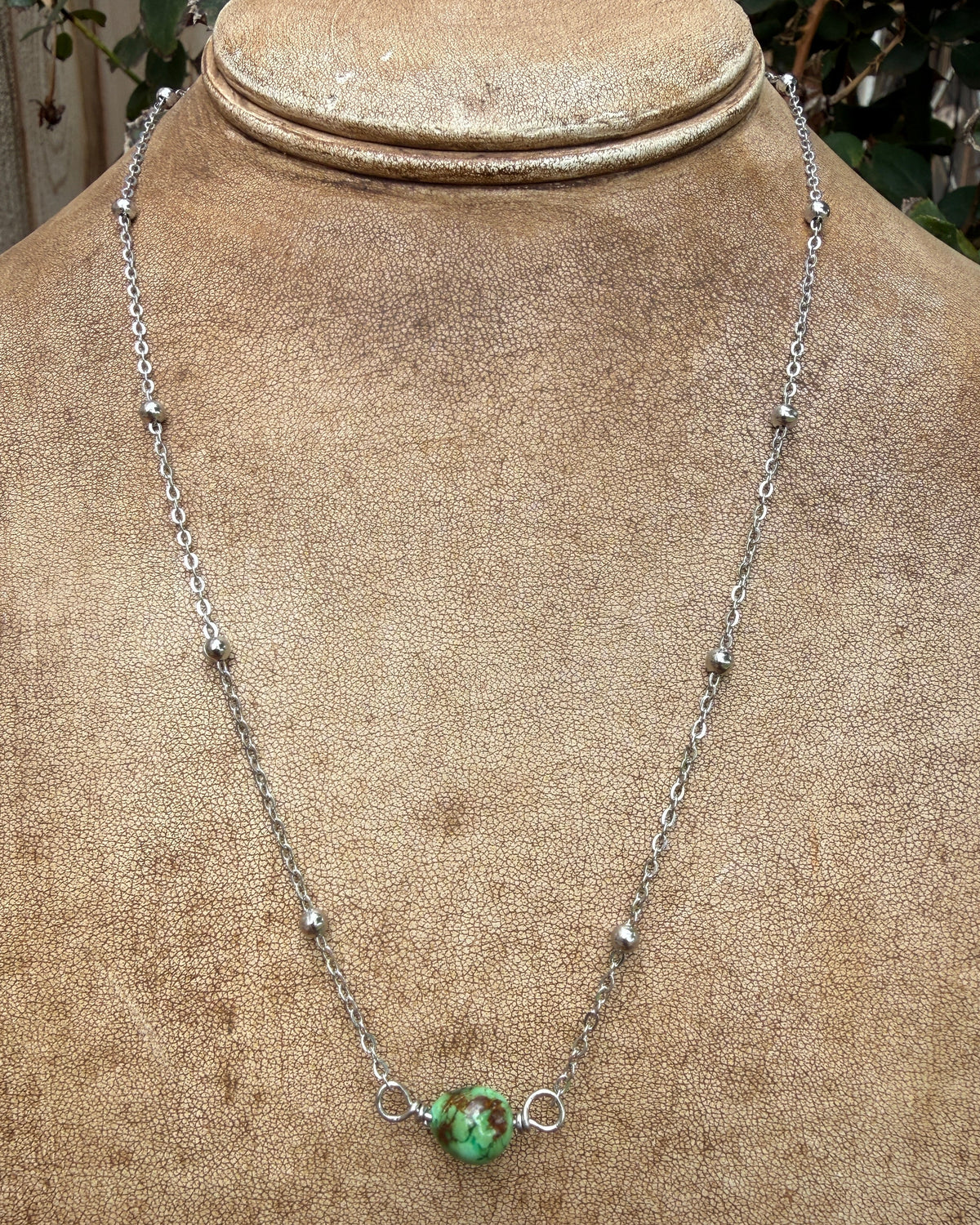 A silver chain necklace with small metallic beads and a central green turquoise bead pendant, displayed on a tan fabric-covered jewelry bust.