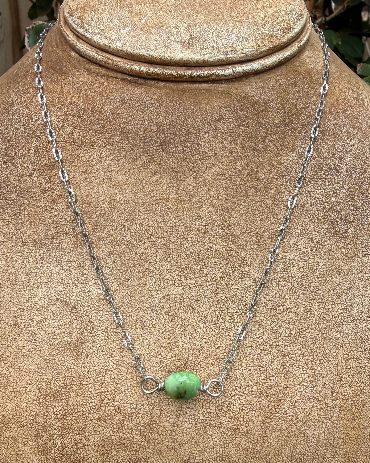 A silver chain necklace with a small green turquoise bead pendant, displayed on a tan fabric jewelry bust.