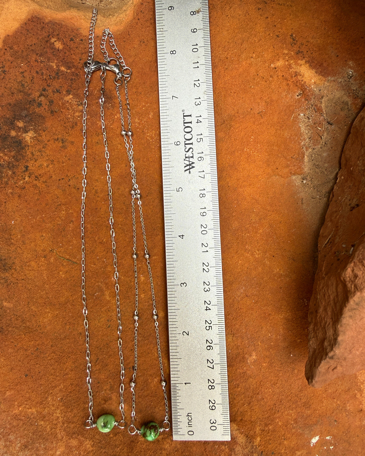 A silver-toned chain necklace with two small green turquoise  bead pendants, shown next to a metal ruler on a textured orange-brown surface.