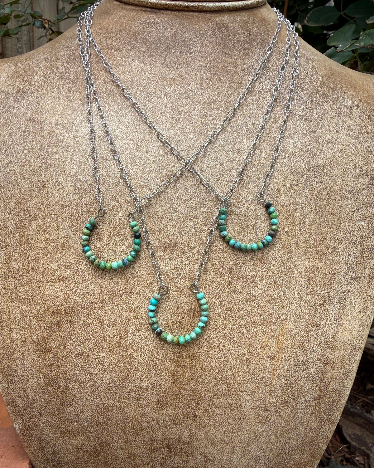 Three layered silver chain necklaces with small turquoise bead horseshoe pendants, displayed on a textured tan jewelry bust.