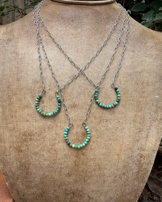 Three layered silver chain necklaces with small turquoise bead horseshoe pendants, displayed on a textured tan jewelry bust.