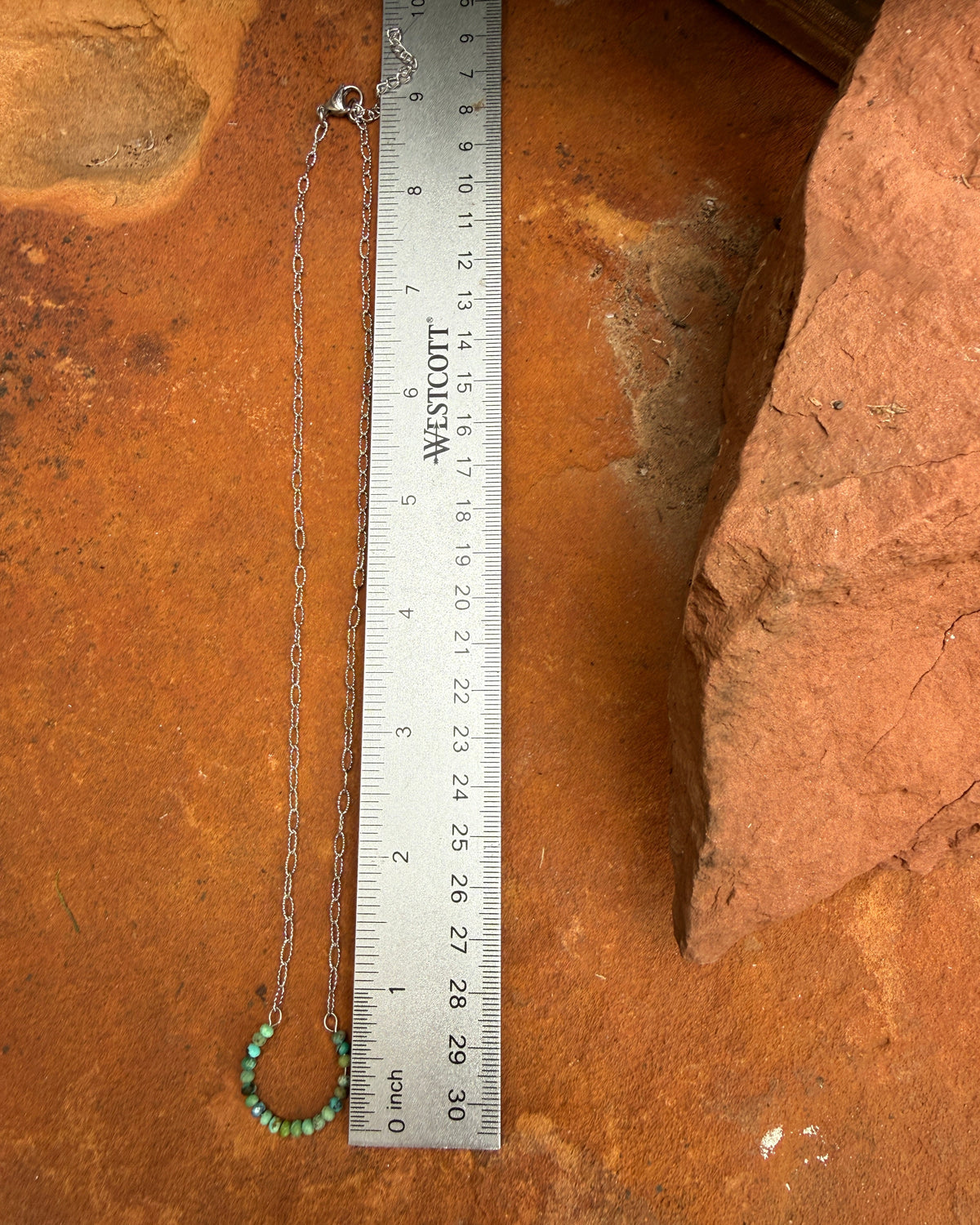 A silver chain necklace with a small horseshoe pendant made of turquoise beads, shown next to a metal ruler for scale on a textured orange-brown rock surface.