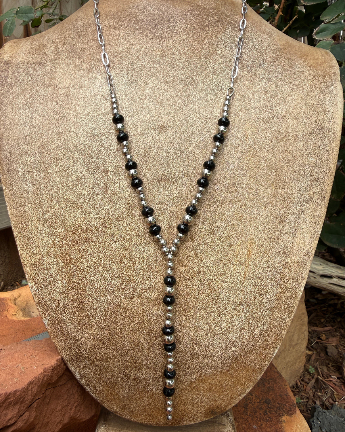 A silver and black beaded Y-necklace displayed on a tan fabric jewelry bust, with an outdoor garden background visible behind the bust.