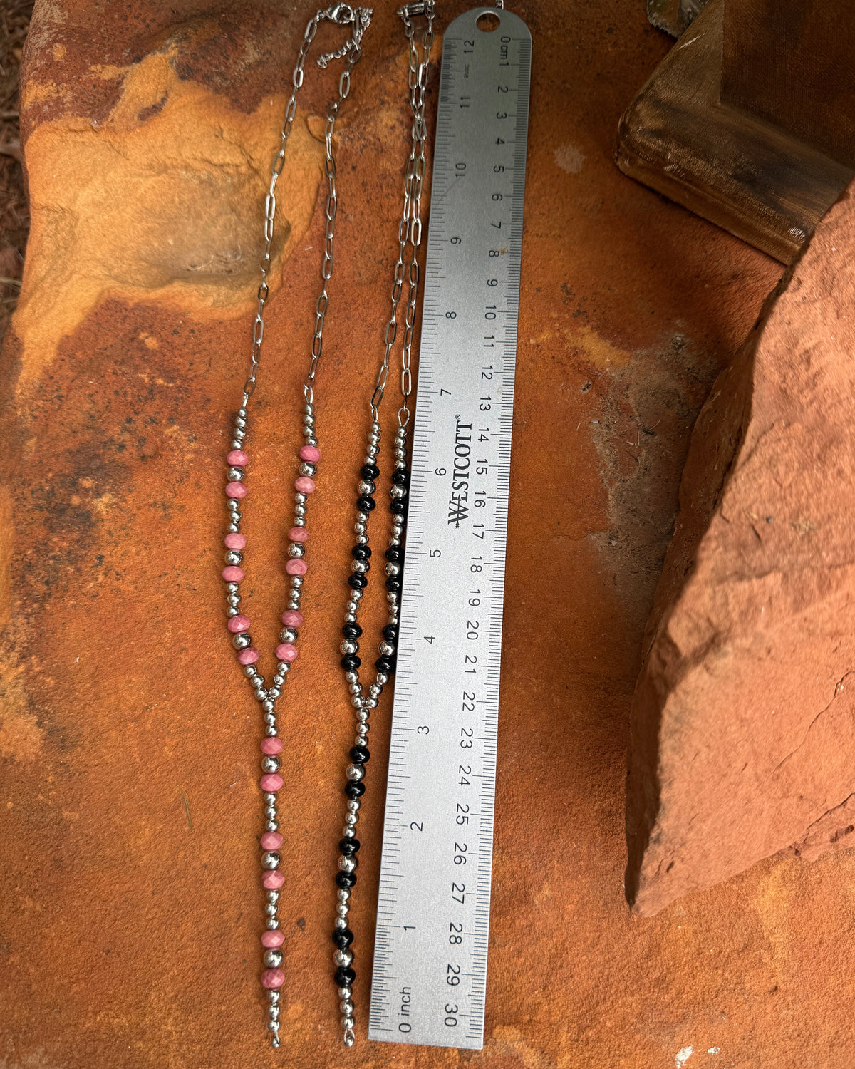 A beaded chain necklace with pink and black beads and silver-toned metal links, laid out on a rusted orange metal surface next to a metal ruler for scale.