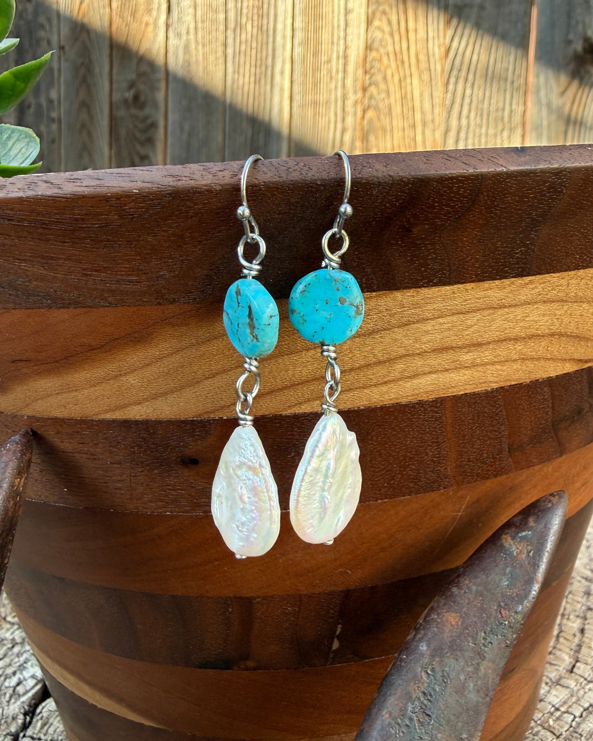 A pair of handmade dangle earrings displayed on a wooden bowl. The earrings feature round turquoise-blue beads on the top and teardrop-shaped white freshwater pearl drops on the bottom, suspended from silver hooks.