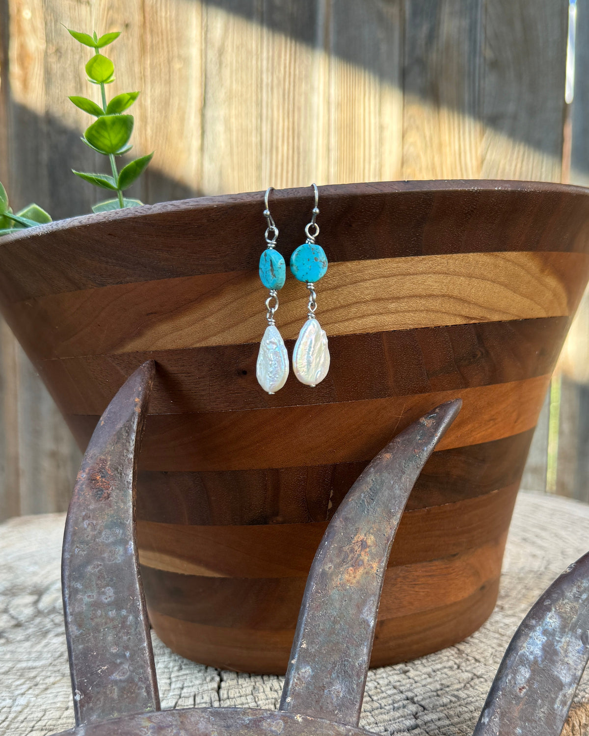 Turquoise and white freshwater pearl teardrop dangle earrings displayed on a wooden bowl, set on a rustic metal wheel prop in front of a wooden fence.