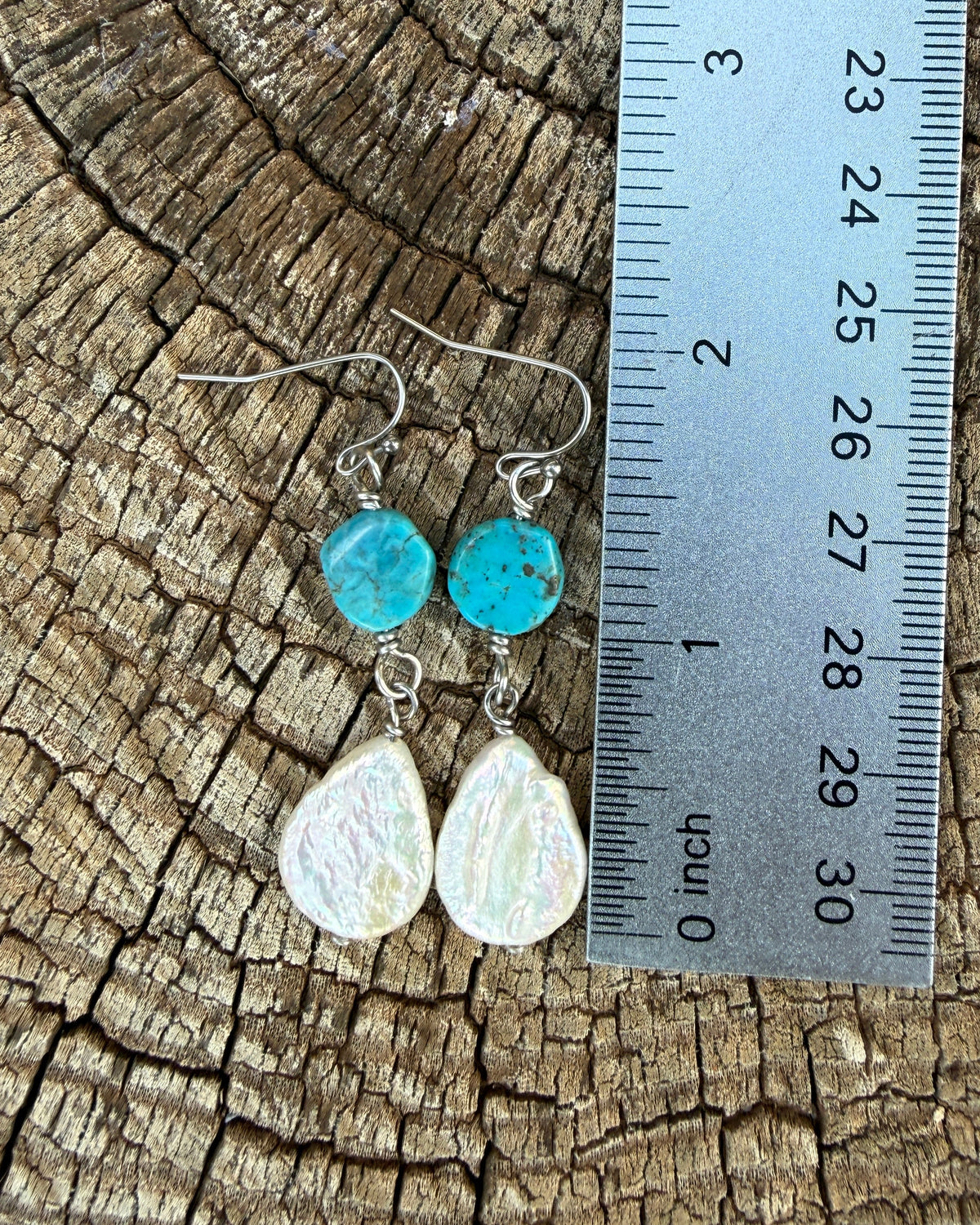 A pair of handmade dangle earrings displayed next to a metal ruler on a textured wood background. The earrings feature turquoise blue beads and white teardrop-shaped freshwater pearl drops on silver wire hooks.