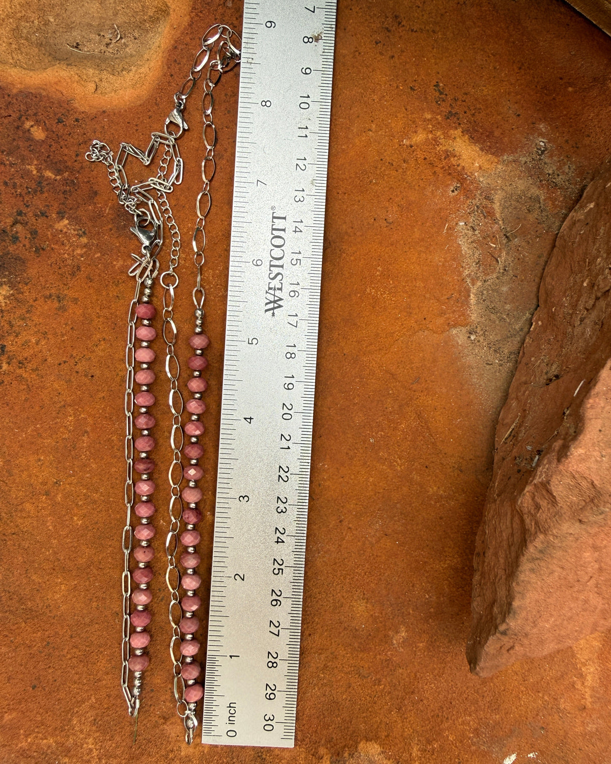 A stainless steel chain necklace with pink faceted rhodonite beads and a matching chain laid out on a rusted metal surface next to a white measuring ruler for scale.