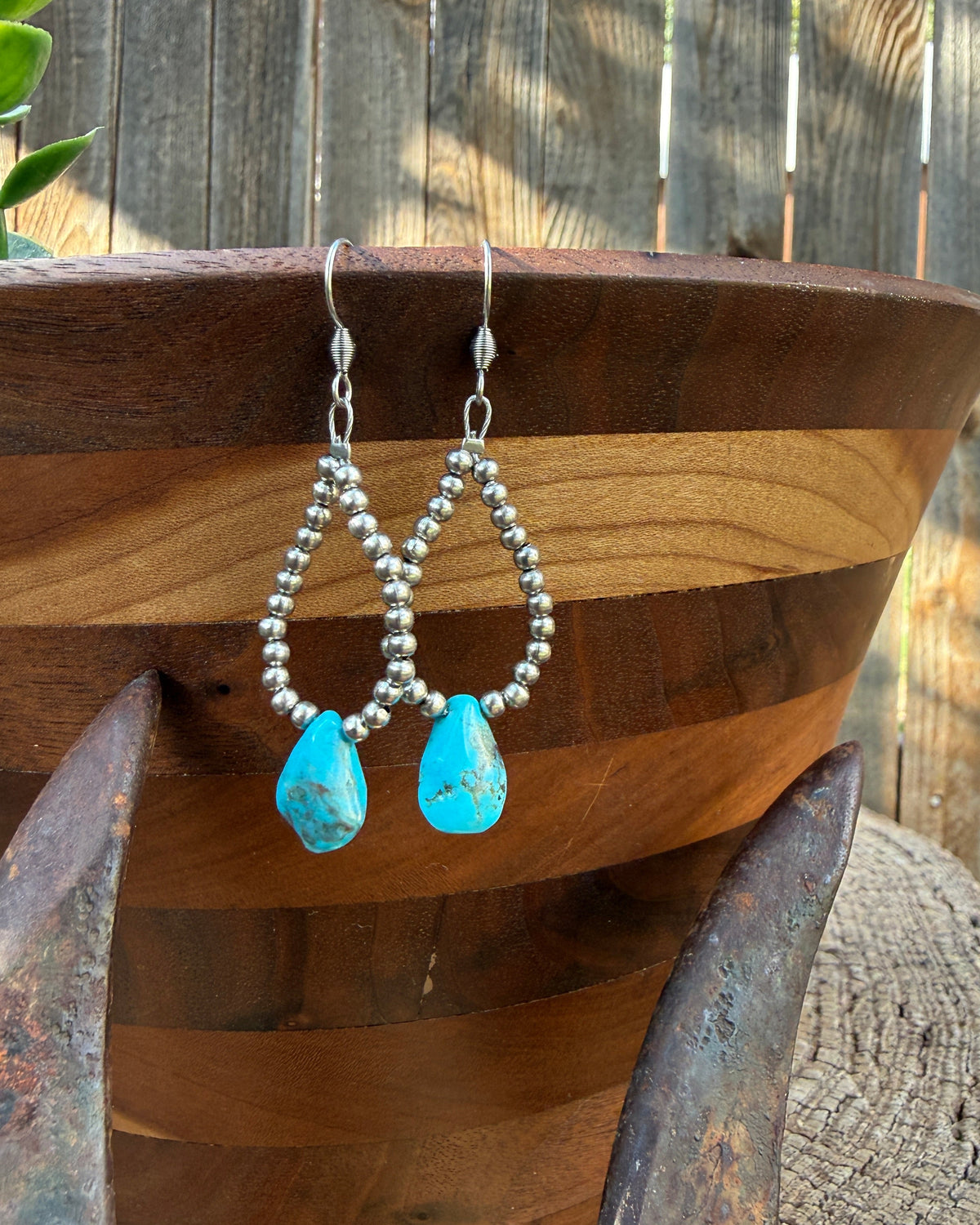 A pair of silver beaded dangle earrings with turquoise blue teardrop beads, hanging from a wooden bowl outdoors against a wooden fence.