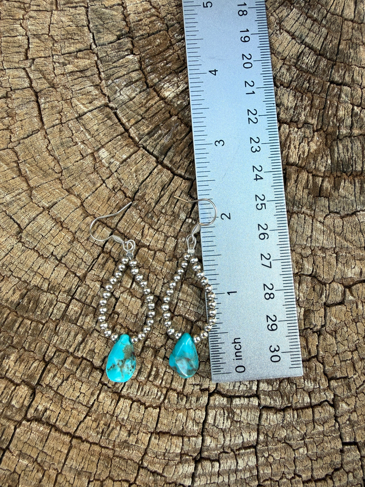 A pair of turquoise and silver dangle earrings displayed on a weathered wood log next to a metal ruler for scale.