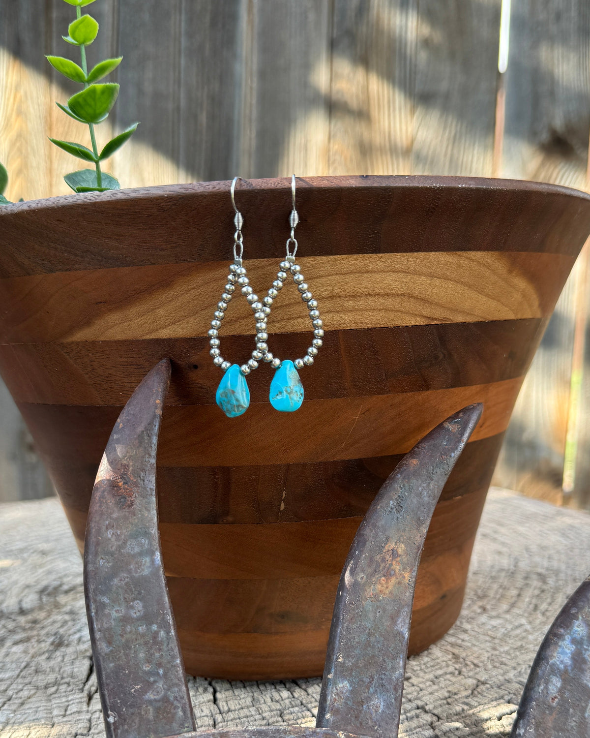 Silver beaded dangle earrings with bright blue teardrop charms hanging from a wooden planter on a rustic metal wheel in front of a wooden fence.
