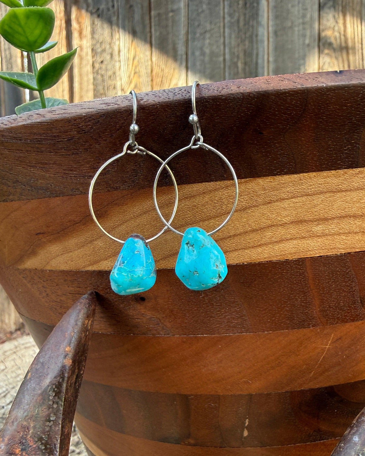 Silver hoop earrings with turquoise stone drops, displayed on a wooden bowl outdoors.