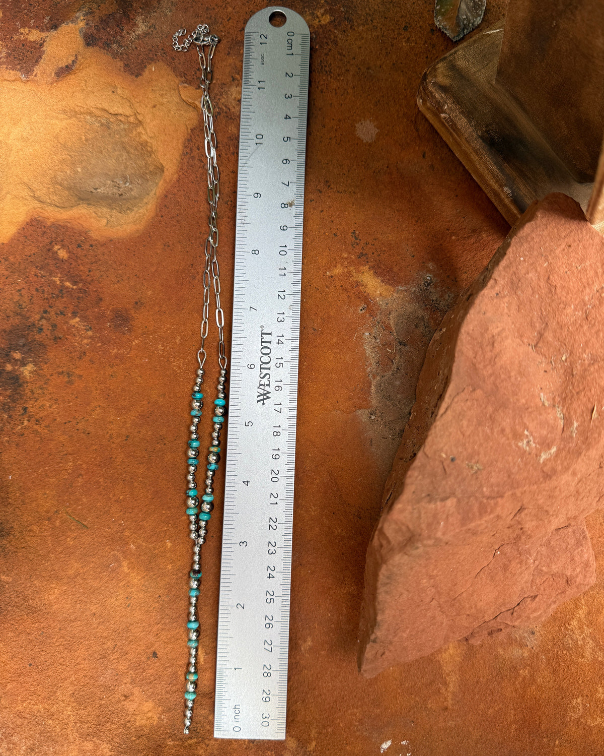 A beaded chain necklace with turquoise and brown beads laid out on a rust-colored surface next to a metal ruler for scale.