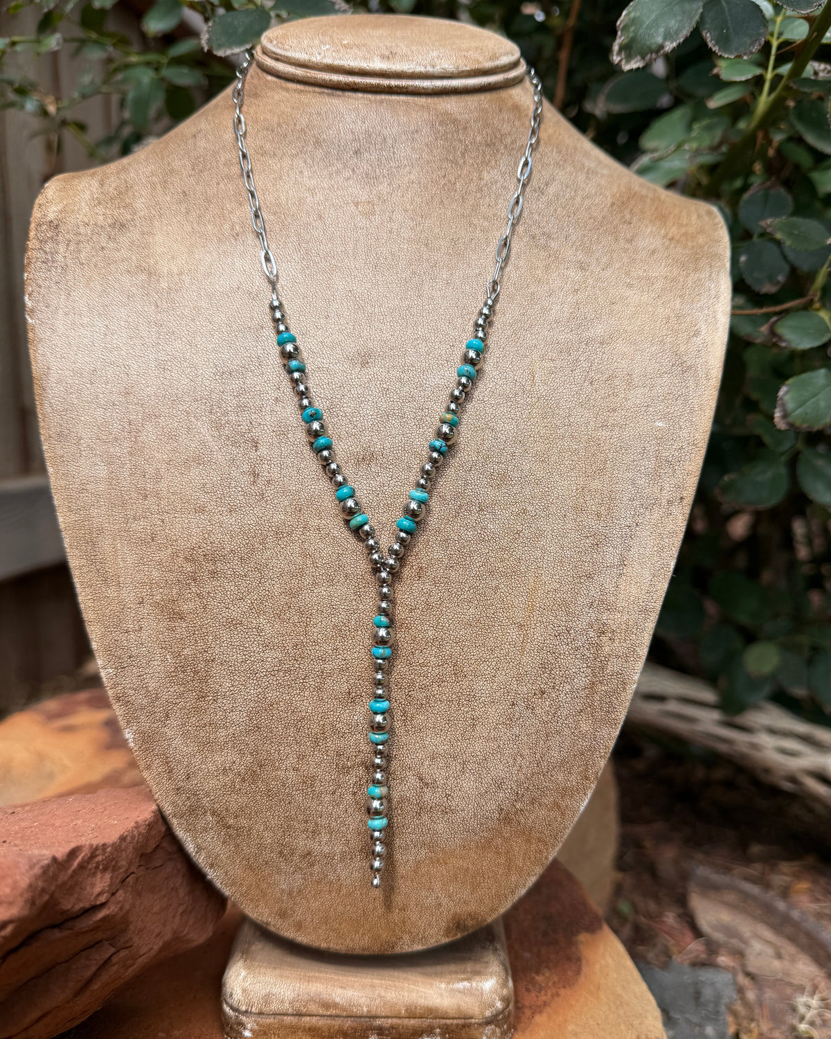 A silver-toned Y-necklace with turquoise and stainless steel beads, displayed on a textured tan jewelry bust with a rustic outdoor background of green leaves and reddish-brown stones.